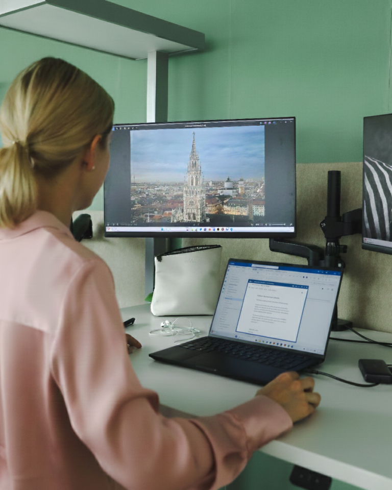 Blonde Frau steht vor zwei Bildschirmen, auf dem einen ist die Stadt München zu sehen, auf dem anderen ein E-Mail Programm